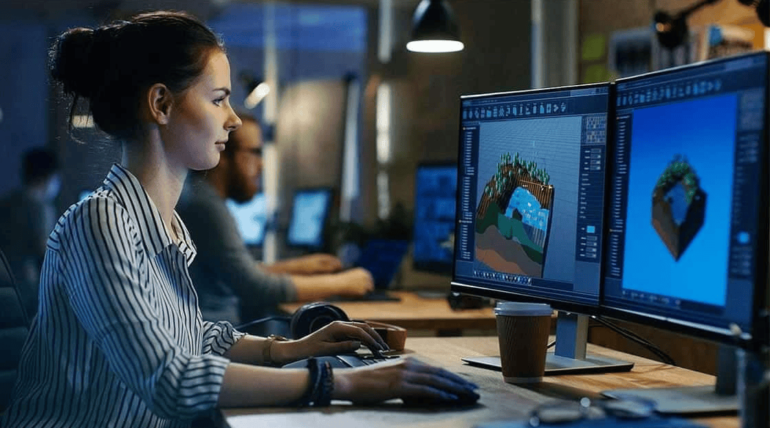 A person working on 3D modeling on two computer monitors, in an office setting with other individuals in the background, engaged in high-tech manufacturing and virtual build projects.