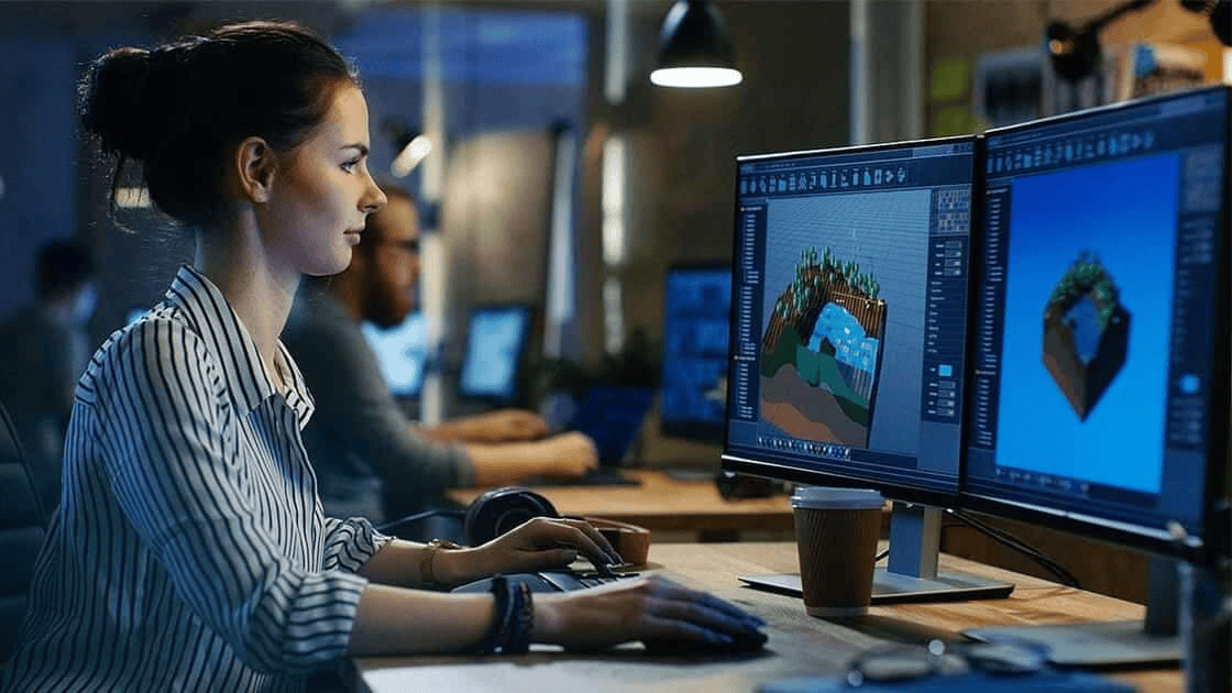 A person working on 3D modeling on two computer monitors, in an office setting with other individuals in the background, engaged in high-tech manufacturing and virtual build projects.